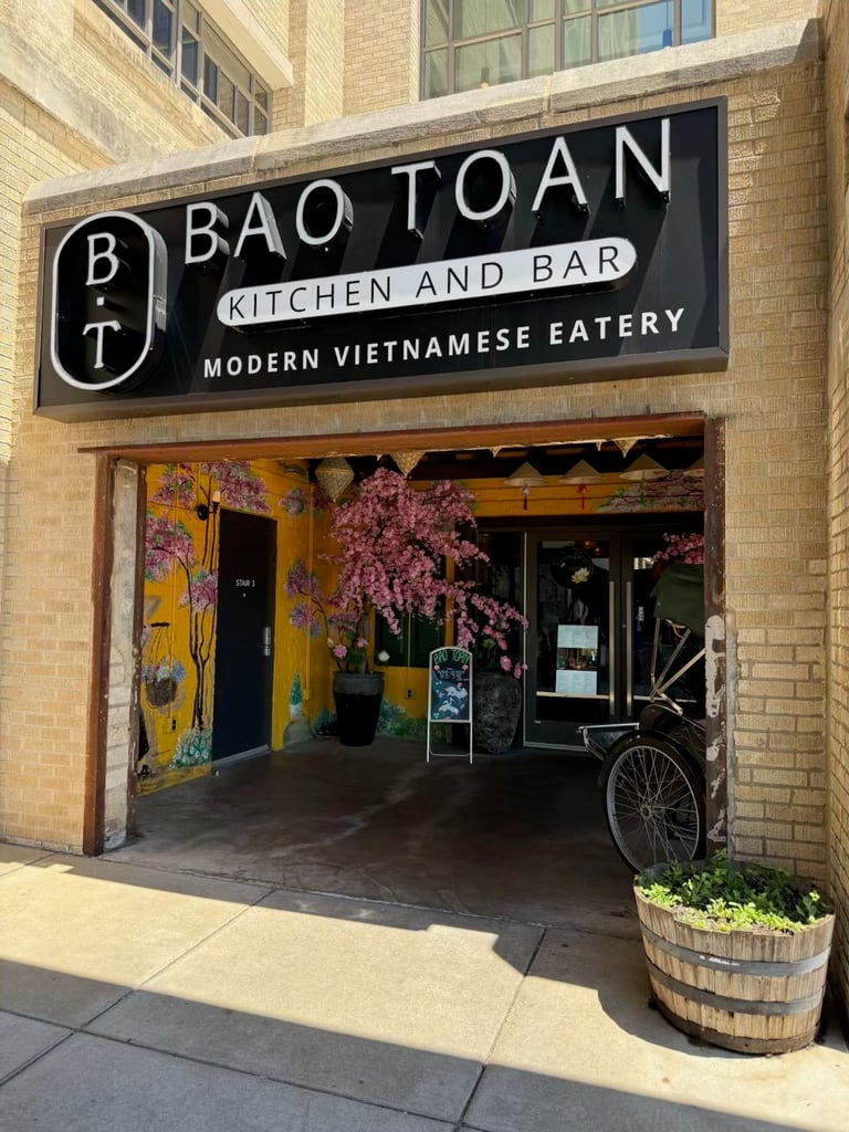 Storefront of Bao Toan Kitchen and Bar, a modern Vietnamese eatery with black signage, colorful yellow door, and flowering plants decorating the entrance on a brick building